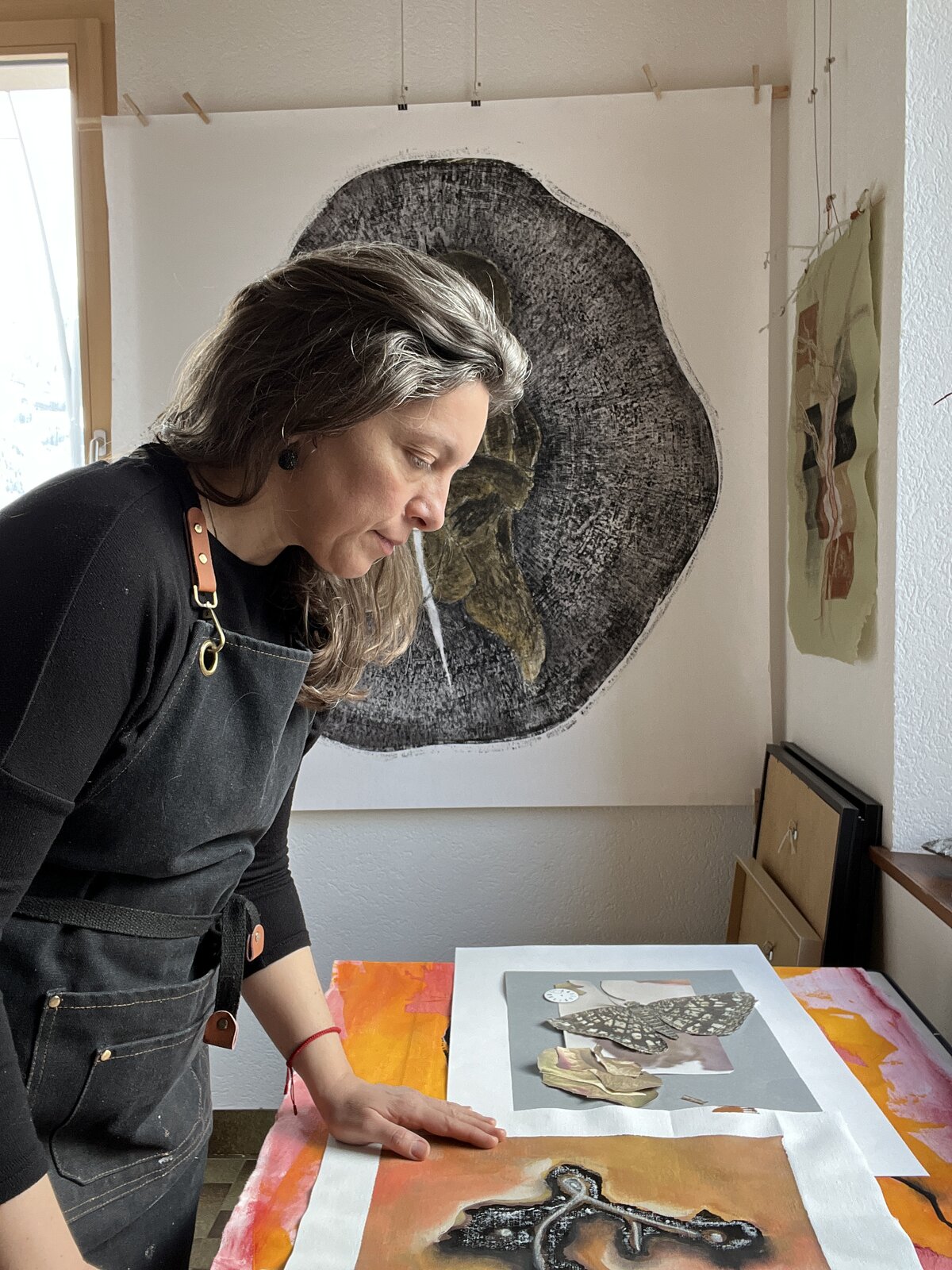 Brindusa Burrows in her studio, examining a print from the (H)Être series alongside collage materials on the worktable.